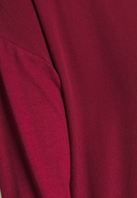 Zbliżenie na bordową tkaninę z widocznymi liniami przeszyć oraz gładką teksturą, ukazującą fałdy i miękkie drapowanie.