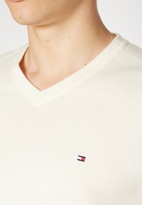 Maglione crema a scollo a V realizzato in tessuto morbido, con un piccolo logo blu, bianco e rosso sul lato sinistro del petto. Texture liscia e design aderente.