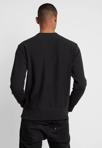 Uomo dai capelli corti che indossa una felpa nera a maniche lunghe e jeans neri, in piedi con la schiena rivolta verso uno sfondo chiaro e semplice.