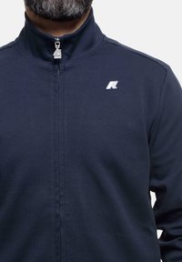 Giacca a zip della Marina con colletto alto, texture a costine e logo ricamato sul lato sinistro del petto. Presenta una zip argentata e un design slanciato.
