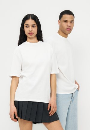 Jeune femme portant une chemise blanche à manches courtes et une jupe plissée noire, debout à côté d'un jeune homme en chemise blanche à manches longues et jean bleu clair.