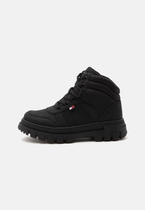 Botte montante noire avec semelle épaisse à crampons, col de cheville rembourré, laçage à l’avant et petit détail de drapeau rouge, blanc et bleu sur le côté.