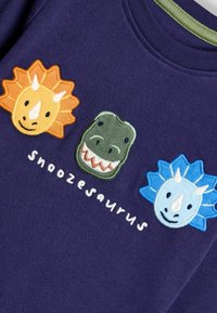 Tummansininen lyhythihainen toppi, jossa on kolme brodeerattua dinosauruksen päätä oranssina, vihreänä ja sinisenä, ja tekstinä "Snoozesaurus" alla.