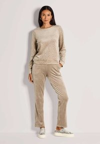 Vrouw die tegen een lichte muur staat, gekleed in beige velours broeken en een bijpassend langemouwen leopoordenprinttop met witte platform sneakers.