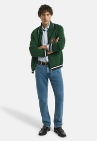 Mann mit lockigem Haar trägt eine grüne Zip-Up-Jacke, ein kariertes Hemd, eine blaue Jeans, einen schwarzen Gürtel und schwarze Schuhe und steht mit verschränkten Armen.