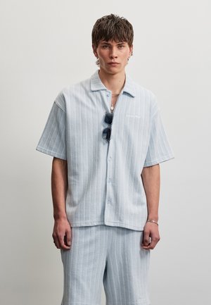 Junger Mann, der ein strukturiertes hellblaues kurzärmeliges Hemd und passende Shorts trägt, mit einer Sonnenbrille, die am Hemdkragen hängt.