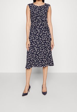 Vrouw draagt een mouwloze marineblauwe midi-jurk met bloemmotief en geplooide details bij de taille, gecombineerd met marineblauwe pumps met puntige neus.