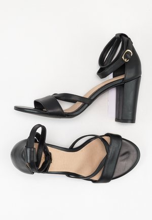 Para czarnych sandałów na wysokim obcasie z paskami na kostkę i złotymi klamrami, jeden z widokiem z boku, a drugi z widokiem od góry na białym tle.