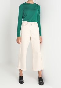 Grön långärmad stickad topp i kombination med vita vida byxor med fransade kanter och svarta skor med dekorativa detaljer.