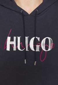 Gros plan sur un sweat à capuche bleu marine avec le texte "HUGO" en blanc et un accent en cursive rose, avec des cordons noirs et des embouts métalliques au niveau de l'encolure.