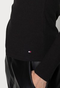 Zwarte geribbelde lange mouw top met een klein logo-label met rode, witte en blauwe accenten; gecombineerd met zwarte leren broek.