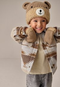 Veste marron à motifs avec boutons-pression, bonnet beige en tricot avec oreilles, et gants assortis. Une chemise claire et un jean gris complètent la tenue.