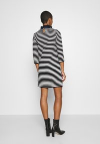 Raidoitu mekko, jossa on kolme neljännestä hihoista, tummansininen ja valkoinen kuosi, röyhelökaulus, avainaukkotakaosa, esitetty mustilla nilkkasaappailla.