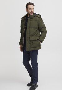 Grön vadderad jacka med huva, dragkedja och stora framlommor. Bärs över en svart topp och kombineras med marinblå byxor och svarta stövlar.