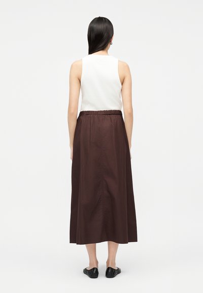 Žena s dlhými tmavými vlasmi, oblečená v bielom topu bez rukávov, hnedej midi sukni s elastickým pásom a čiernych slip-on topánkach, stojaca chrbtom k nám.