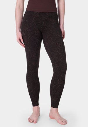 Vrouw draagt donkerbruine legging met subtiele luipaardprint, staat blootsvoets tegen een effen lichte achtergrond.