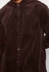 Hnědá košile z manšestru s límečkem na knoflíky, svislou vroubkovanou texturou a černými knoflíky vpředu.