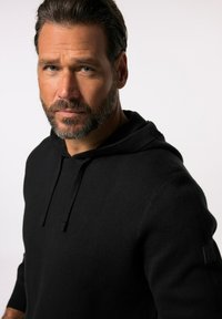 Schwarzer Hoodie aus weichem Material mit Kordelzug, ausgestattet mit einem lederähnlichen Logo-Patch am linken Ärmel und gerippten Bündchen.