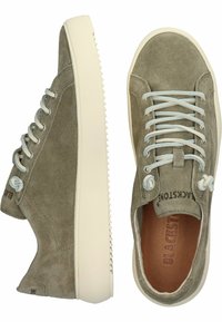 Šviesiai alyvuogių spalvos zomšos sportbačiai su plokščiomis šviesiai mėlynomis raištelėmis, balta gumine padu ir iškilniu logotipu ant išorinės pusės.