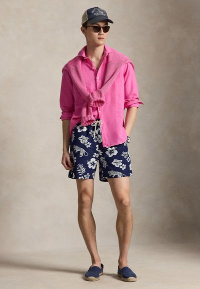 Hombre con camisa rosa y un suéter a juego sobre los hombros, pantalones cortos azul marino con estampado floral, zapatos náuticos azul marino, gafas de sol y una gorra de béisbol.