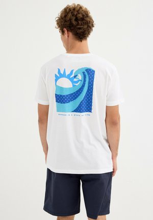 Hombre vistiendo camiseta blanca con gráfico estilizado de sol y ola azul y pantalones cortos azul marino, de pie mirando hacia atrás sobre fondo liso.