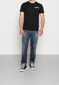 Man som bär en svart ellesse t-shirt, urtvättade blå jeans med uppvikta ben, och svarta högstövlar som står mot en vit vägg.