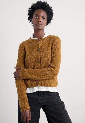 Mustard-colored knitted cardigan with a round neckline, two brown buttons, ribbed cuffs, and a cropped design, layered over a white shirt.