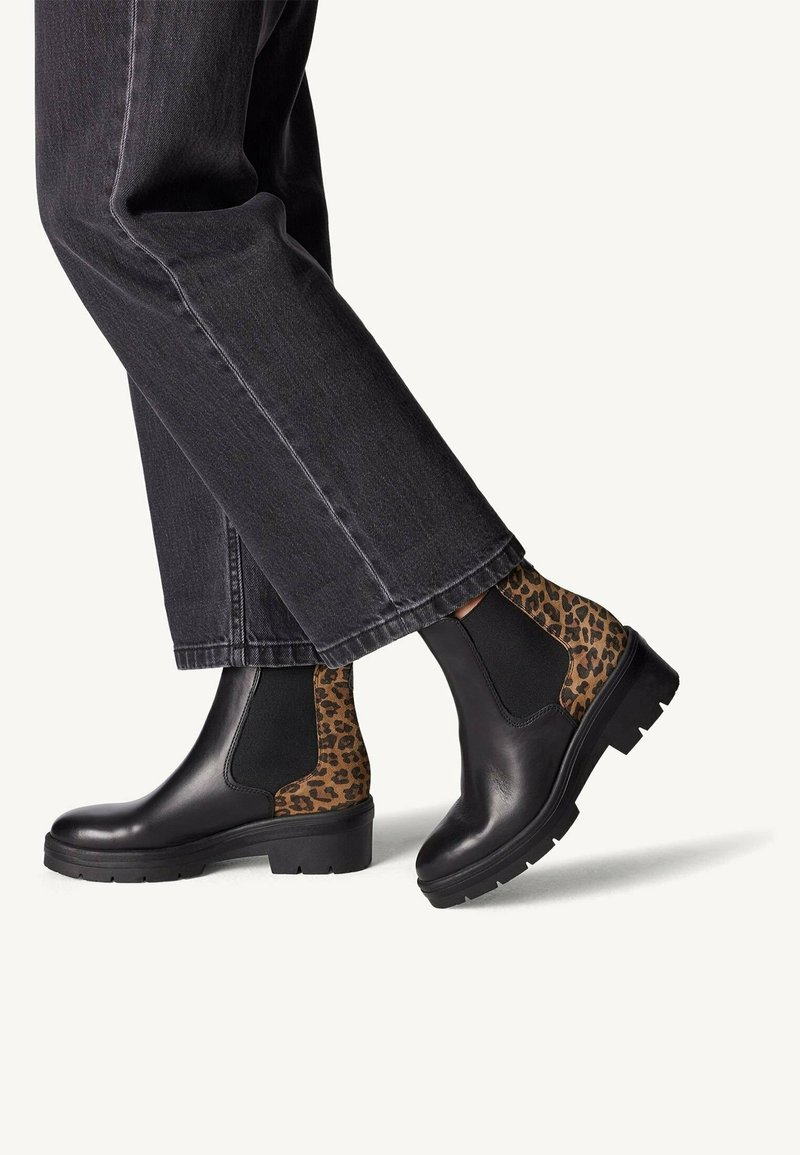 Personne portant des bottines Chelsea en cuir noir avec des panneaux élastiques à imprimé léopard et un jean noir sur fond blanc.