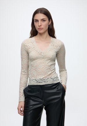Giovane donna con lunghi capelli castani che indossa un top beige in pizzo a maniche lunghe e pantaloni di pelle neri, in piedi davanti a uno sfondo bianco.