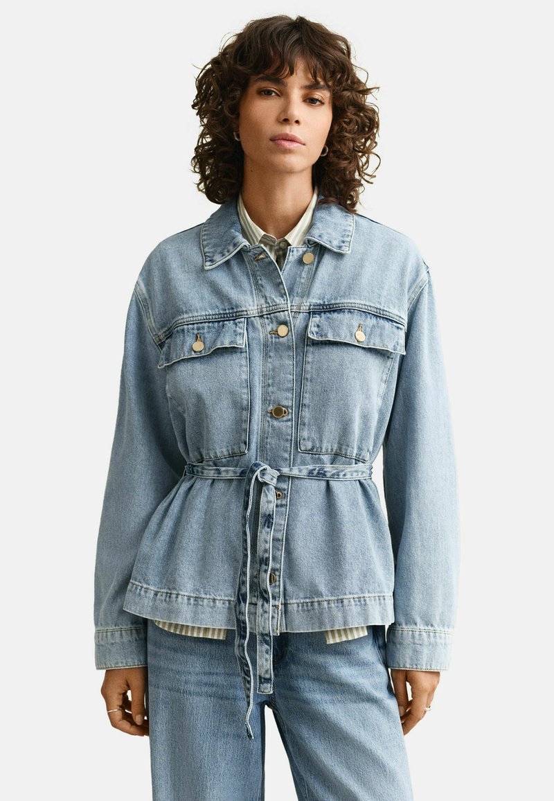 Femme aux cheveux bouclés portant une veste en jean bleu clair ceinturée sur une chemise rayée et un jean assorti, faisant face.