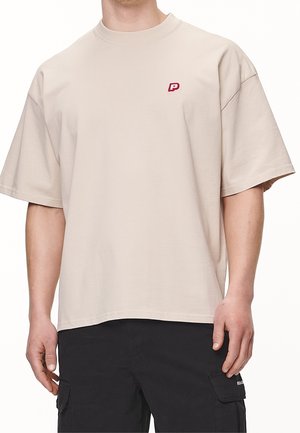 T-shirt in cotone beige con collo rotondo e maniche corte, con un piccolo logo rosso "P" sul lato sinistro del petto. Vestibilità ampia e rilassata.