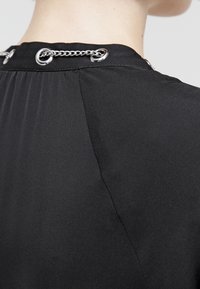 Vêtement noir à la texture lisse, doté d'un col rond et d'un détail en chaîne argentée au dos, mettant en avant un design minimaliste.