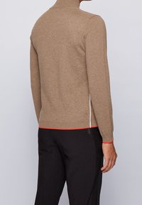 Man som bär en ljusbrun långärmad tröja med röd kant vid fållen och svarta byxor med subtila strukturpaneler på sidorna.