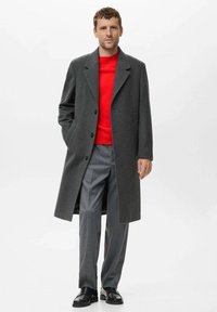 Cappotto in lana grigio carbone con colletto a revers, chiusura con bottoni e tasche laterali, indossato sopra un maglione rosso brillante e pantaloni grigi.