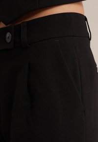 Črne krojene hlače iz teksturiranega materiala. Značilnosti vključujejo raven pas z gumbom in stranske gube za dodatno podrobnost.