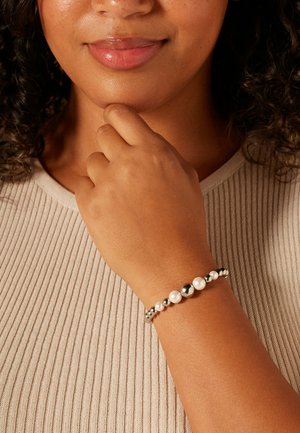 Zilveren en parelarmband met afwisselend glanzend zilver en witte parels op een slank bandje, gedragen aan een pols tegen een gestructureerde beige achtergrond.