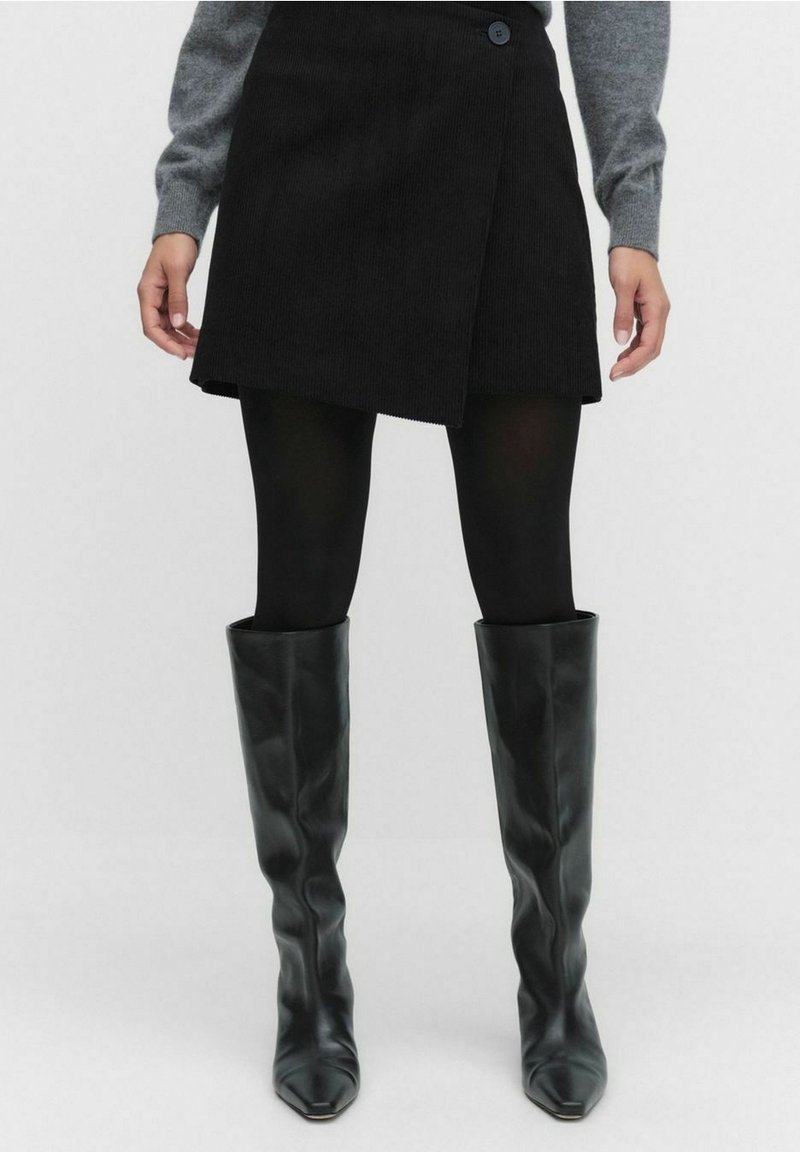 Personne portant un pull gris, une mini-jupe portefeuille noire, des collants noirs et des bottes noires en cuir jusqu’aux genoux, debout devant un fond uni.