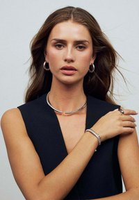 Smycken i silver med ett krökt halsband, örhängen i form av ringar, ett armband med texturerad metalldesign och en metallring på en modell.