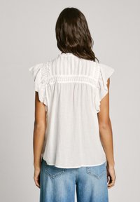 Weißes ärmelloses Oberteil mit Rüschenschultern, das auf der Rückseite mit Raffungen versehen ist. Kombiniert mit hellblauen, hochgeschnittenen Jeans.