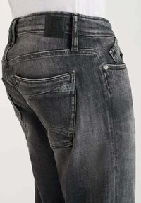 Jeans en denim gris avec une finition délavée, comportant cinq poches, des coutures contrastées et un patch en cuir avec logo sur la ceinture.