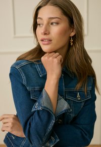 Nuori nainen, jolla on pitkät ruskeat hiukset, kullanväriset korvakorut ja tumma farkkutakki, leuka nyrkin päällä ja katse sivulle.