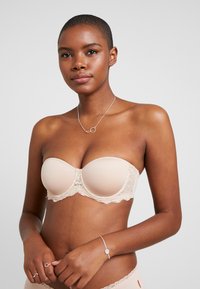 Beige bustier uden stropper med blondekant og formfaste skåle, der har glat stof og en struktureret form. Model, der viser produktet.