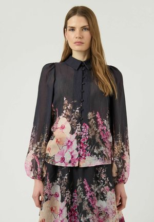 Žena s dlouhými vlasy, oblečená v černém průsvitném topu a sukni s barevnými růžovými a krémovými květinovými vzory a knoflíky potaženými látkou.