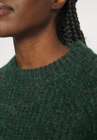 Gros plan sur une personne portant un pull en laine tricotée vert foncé texturé, montrant le cou, le menton et les cheveux tressés.