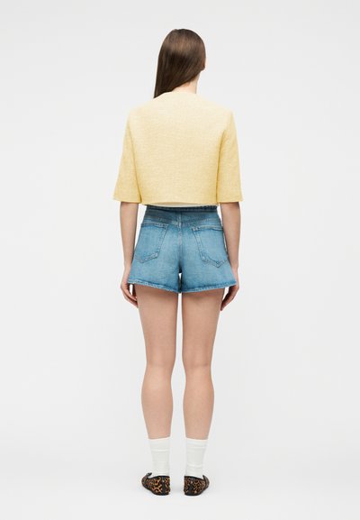 Femme aux longs cheveux bruns portant un haut jaune court, un short en jean bleu taille haute, des chaussettes blanches et des ballerines à motif léopard, vue de dos.