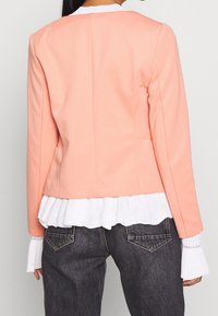 Blazer ajusté de couleur pêche avec une coupe structurée, présentant un ourlet plissé blanc visible en dessous, associé à un pantalon en denim foncé.