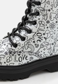 Stivale alla caviglia bianco in pelle decorato con motivi neri a doodle, tra cui cuori e stelle, dotato di lacci neri e una suola in gomma resistente.