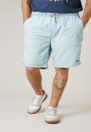 Homme portant un short bleu clair avec poches avant, une chemise marine, des baskets blanches et des tatouages visibles sur les jambes et les avant-bras.