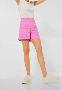 Person klädd i höga rosa shorts, vit t-shirt och vita platåskor som står mot en enkel bakgrund.