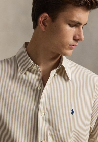 Smėlio spalvos ir baltos spalvos dryžuota palaidinė su sagomis, su smailiu apykakliu ir dviem sagomis priekyje, kairėje krūtinės pusėje siuvinėtas mėlynas logotipas.